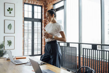 Weibliche Geschäftsfrau mit Kaffeetasse, die auf einen Laptop schaut, während sie im Büro steht - VPIF05007