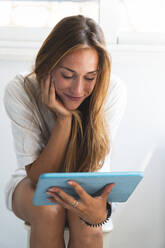Frau mit Hand am Kinn, die auf einem Hocker sitzend ein digitales Tablet betrachtet - JAQF00750