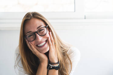 Happy woman wearing eyeglasses sitting with head in hand at home - JAQF00753