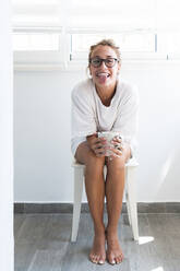 Young woman with coffee mug sticking out tongue while sitting on stool at home - JAQF00759