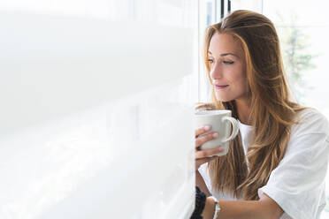 Schöne junge Frau mit Tasse schaut durch das Fenster zu Hause - JAQF00761