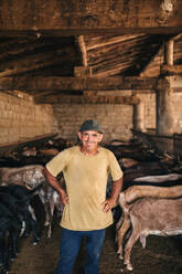 Happy male goat herder standing with hand on hip in shed - GRCF00891