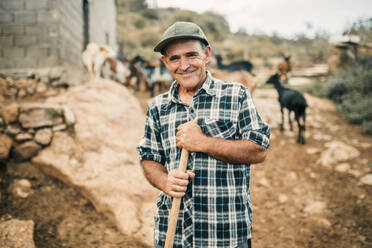 Lächelnder männlicher Ziegenhirte, der einen Besen hält, während er auf einem Bauernhof steht - GRCF00902