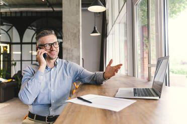 Smiling mature businessman gesturing while talking on mobile phone in office - OIPF01094