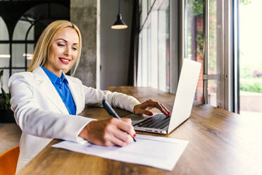 Smiling businesswoman writing on paper while sitting with laptop at desk in office - OIPF01097