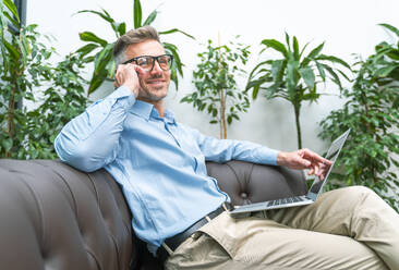 Lächelnder Geschäftsmann mit Laptop, der mit seinem Smartphone im Büro spricht - OIPF01119