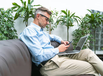 Männlicher Geschäftsmann sitzt mit Laptop und benutzt ein Mobiltelefon im Büro - OIPF01120