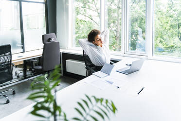 Junge Geschäftsfrau mit Händen hinter dem Kopf sitzt am Schreibtisch im Büro - OIPF01138