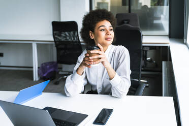 Businesswoman holding disposable coffee cup while sitting with laptop in office - OIPF01141