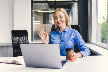 Lächelnde blonde Geschäftsfrau winkt mit der Hand während eines Videoanrufs über einen Laptop im Büro - OIPF01148