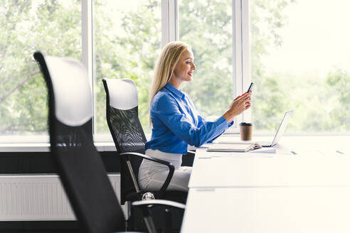 Blonde Geschäftsfrau, die mit ihrem Smartphone Textnachrichten verschickt, während sie am Schreibtisch im Büro sitzt - OIPF01151