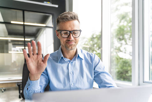 Männlicher Geschäftsmann winkt mit der Hand während eines Videoanrufs über einen Laptop im Büro - OIPF01167