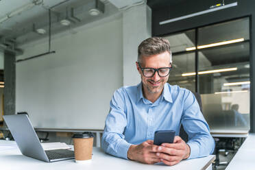 Smiling businessman using mobile phone at desk in office - OIPF01170