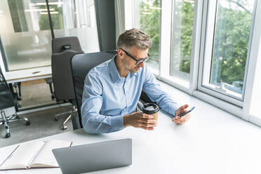 Businessman with coffee cup using mobile phone at desk in office - OIPF01173