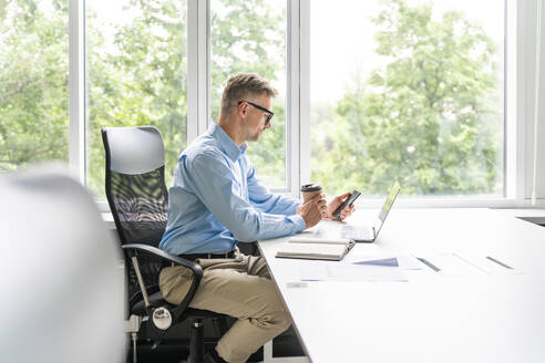 Reifer Geschäftsmann, der ein Mobiltelefon benutzt, während er im Büro sitzt - OIPF01176