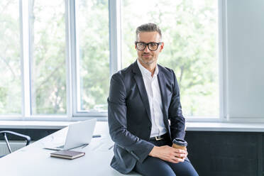 Älterer Geschäftsmann mit Brille sitzt auf dem Schreibtisch im Büro - OIPF01197