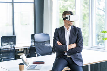 Male business professional wearing virtual reality headset while sitting with arms crossed in office - OIPF01207