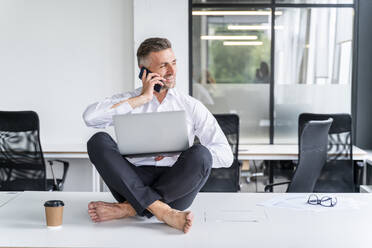 Male business person sitting with laptop while talking on smart phone in office - OIPF01224