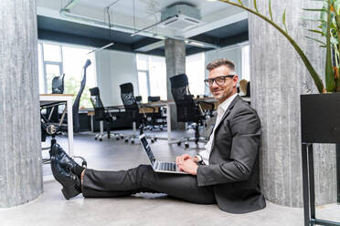 Lächelnder Geschäftsmann mit Laptop auf dem Boden sitzend im Büro - OIPF01226