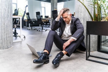 Worried businessman with head in hand sitting on floor in office - OIPF01228