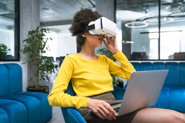 Geschäftsfrau mit Laptop, die ein Virtual-Reality-Headset trägt, während sie in einer Büro-Lobby sitzt - OIPF01272