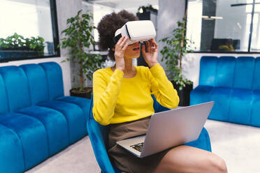 Junge Geschäftsfrau trägt ein Virtual-Reality-Headset, während sie in einer Büro-Lobby sitzt - OIPF01273