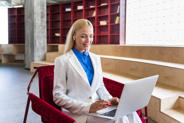 Lächelnde Geschäftsfrau, die einen Laptop benutzt und auf einem Stuhl im Büro sitzt - OIPF01298