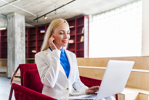 Blonde Geschäftsfrau passt In-Ear-Kopfhörer an, während sie im Büro einen Laptop benutzt - OIPF01299