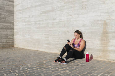 Lächelnde Sportlerin, die ein Mobiltelefon benutzt, während sie an einem sonnigen Tag vor einer Wand sitzt - JRVF01848