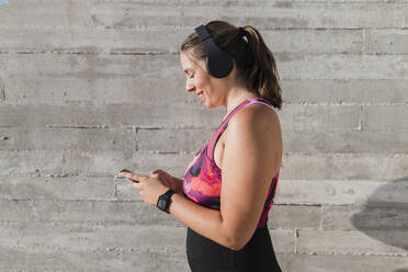 Smiling sportswoman wearing wireless headphones while listening music by wall - JRVF01849