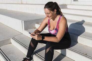 Lächelnde Sportlerin, die auf einer Treppe sitzend eine Textnachricht über ihr Smartphone verschickt - JRVF01855