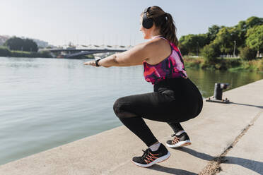 Young sportswoman listening music through wireless headphones while exercising at riverbank - JRVF01865