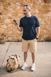 Mann mit Hand in der Tasche, der sein Smartphone benutzt, während er vor einer Wand steht - GRCF00906