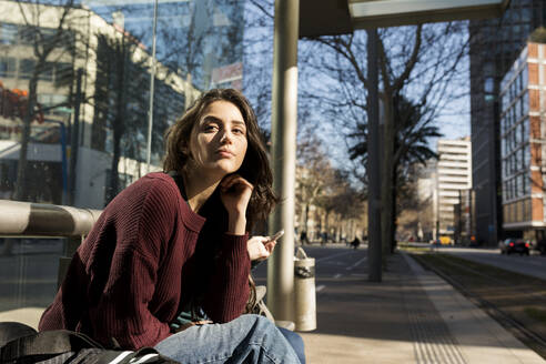 Junge Frau wartet auf Straßenbahn und sitzt auf einer Bank in der Stadt - VABF04317