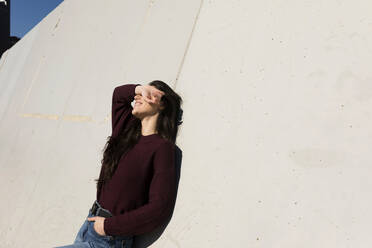 Frau mit Händen in den Hosentaschen, die ihre Augen abschirmt, während sie sich an einer Wand an einem sonnigen Tag anlehnt - VABF04334