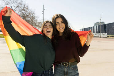 Glückliches lesbisches Paar hält Regenbogenflagge an einem sonnigen Tag - VABF04337