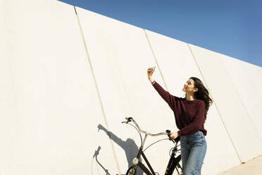 Frau mit Fahrrad macht Selfie mit Smartphone an der Wand - VABF04344