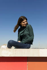 Junge Frau sitzt auf einer Stützmauer an einem sonnigen Tag - VABF04351