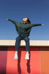 Carefree woman with arms outstretched sitting on retaining wall during sunny day - VABF04352