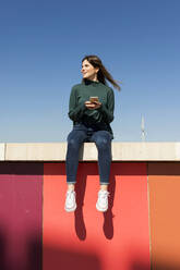 Woman with smart phone sitting on retaining wall - VABF04353