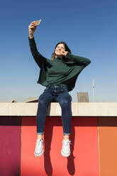 Frau gestikuliert Friedenszeichen, während sie ein Selfie auf einer Stützmauer macht - VABF04354