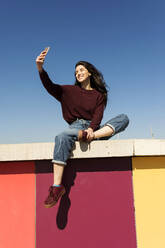 Young woman taking selfie through smart phone on retaining wall - VABF04355