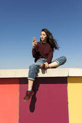 Young woman using smart phone on retaining wall - VABF04356