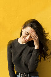 Smiling woman with head in hand in front of yellow wall - VABF04369