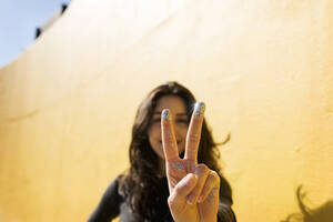 Frau mit Glitzer an den Fingern gestikuliert Friedenszeichen vor einer Wand - VABF04380