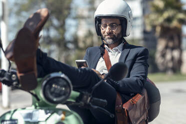 Männlicher Berufstätiger, der ein Mobiltelefon benutzt, während er mit hochgelegten Füßen auf einem Motorroller sitzt - JSRF01641