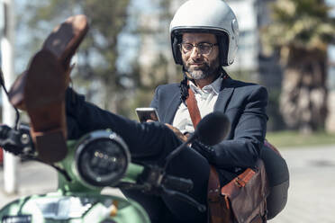 Male professional using mobile phone while sitting with feet up on scooter - JSRF01641