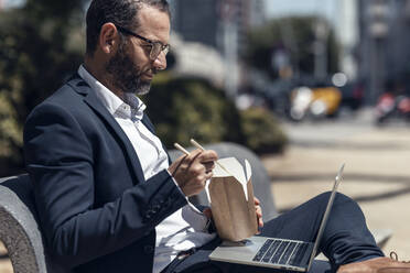 Businessman with laptop eating food while sitting on bench - JSRF01659