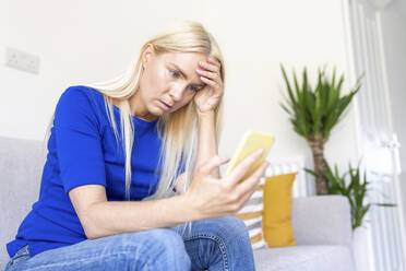 Besorgte Frau mit Kopf in der Hand, die ihr Smartphone hält, während sie zu Hause sitzt - WPEF05281