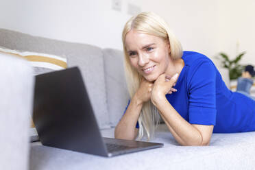 Mature woman with hands on chin looking at laptop while lying at home - WPEF05290